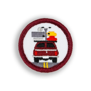 May include: A red embroidered patch with a white background. The patch features a red car with a gray roof rack. On the roof rack are a gray suitcase, a gray camera, and a yellow life preserver. The car is driving on a gray road with yellow lines.
