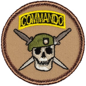May include: A tan and brown embroidered patch with a skull wearing a green beret and crossed daggers. The patch has a yellow tab with the word "COMMANDO" on it.