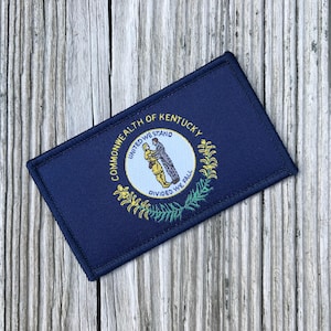 May include: A rectangular patch featuring the Kentucky state flag. The patch is navy blue with a central emblem of a circle containing two figures and the words "UNITED WE STAND, DIVIDED WE FALL." The words "COMMONWEALTH OF KENTUCKY" are arched above the circle.