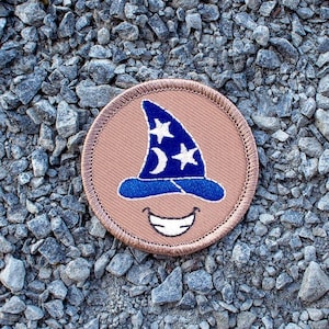 May include: A brown embroidered patch with a blue and white wizard hat and a smiling face. The patch has a tan border.