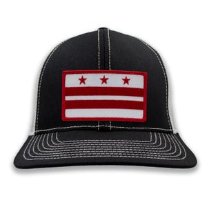 May include: Black and white trucker hat with a rectangular patch featuring a red and white flag design. The flag has three red stripes and three white stripes, with three red stars at the top. The hat has a curved brim and white stitching.