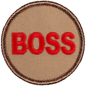 May include: A round patch with the word "BOSS" embroidered in red thread on a tan background. The patch has a brown stitched border. The design is simple and bold, suitable for various applications.