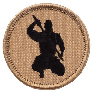May include: Round patch with a tan background and a black silhouette of a ninja in a kneeling position, holding a sword. The patch has a stitched border in a darker tan color.