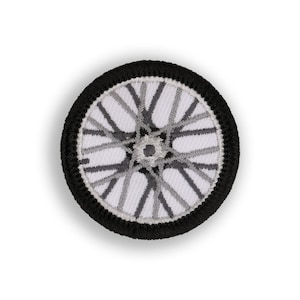 May include: A black and white embroidered patch with a wheel design. The wheel has a white background with black and gray spokes.