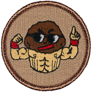 May include: A brown and tan embroidered patch with a cartoon cupcake wearing sunglasses and flexing its muscles. The cupcake is wearing a red wristband on each arm. The patch is circular and has a brown border.
