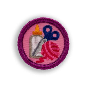 May include: Embroidered patch with a pink background and a purple border. The patch features a glue bottle, blue scissors, and a ball of red yarn. The glue bottle has a yellow cap, and the scissors are open.