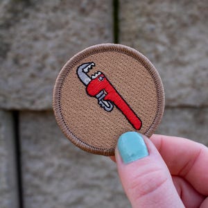 May include: A brown embroidered patch with a red wrench tool. The wrench is a common symbol for a handyman or plumber.