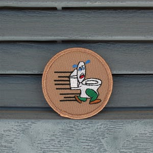 May include: A round, tan embroidered patch featuring a cartoon toilet running with a panicked expression. The toilet is white with green legs and blue sweat drops. Black speed lines suggest movement.