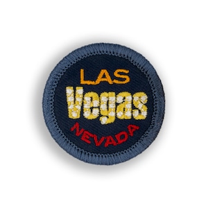 May include: A round, navy blue embroidered patch with the words "LAS VEGAS NEVADA". The word "LAS" is in gold, "VEGAS" is in a checkered pattern of yellow and white, and "NEVADA" is in red.