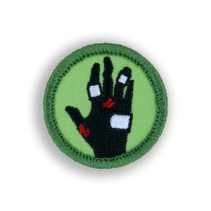 May include: A round, embroidered patch featuring a black hand with red and white accents on a light green background. The patch has a green border. This patch could be used to decorate clothing or accessories.
