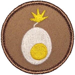 May include: A brown embroidered patch with a white egg with a yellow yolk and a yellow flame on top.