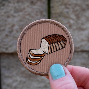 May include: A brown embroidered patch with a white bread loaf and slices of bread. The patch is circular and has a brown border.