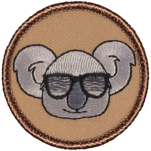 May include: Embroidered patch featuring a cartoon koala wearing sunglasses. The koala is gray with black eyes and a brown nose. The patch is circular and has a brown border.