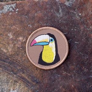 May include: A brown embroidered patch with a toucan with a yellow body, black head, and a blue and pink beak.