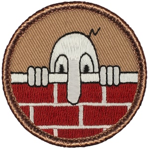 May include: A brown embroidered patch with a white cartoon character peeking over a red brick wall. The character has a large nose and big eyes.