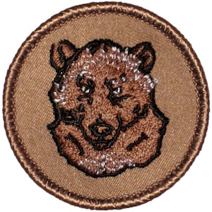 May include: A brown embroidered patch with a brown bear head in the center. The bear has a black nose and eyes. The patch is surrounded by a brown stitched border.