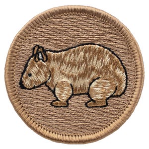 May include: A circular embroidered patch featuring a tan wombat with dark brown outlines. The background of the patch is a textured light brown fabric, and the entire circular edge is finished with a thick, tan stitched border.