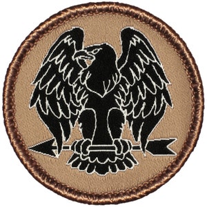May include: A black embroidered patch with a brown border. The patch features a black eagle with wings spread, standing on a spear with an arrow.