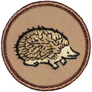 May include: A brown embroidered patch with a beige background featuring a brown and black hedgehog.