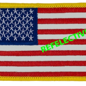 May include: A small, embroidered American flag patch with a yellow border. The text "REFLECTIVE!" is printed in green on the right side of the patch.