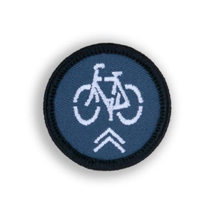 May include: A round, blue embroidered patch with a white bicycle icon and white arrows pointing upwards. The patch has a black border.