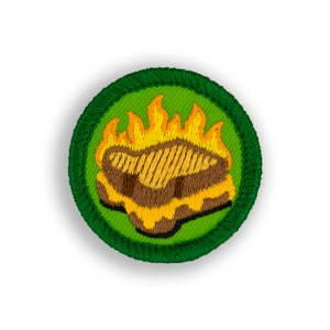 May include: A green and yellow embroidered scout patch with a campfire design. The patch features a brown log with yellow flames.