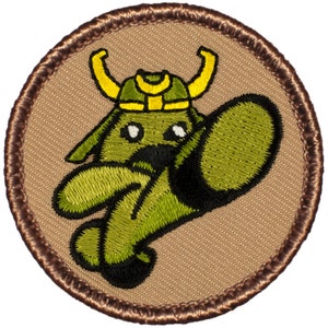 May include: A round, brown embroidered patch with a green cartoon character wearing a yellow helmet with horns. The character is in a fighting pose with one leg raised.