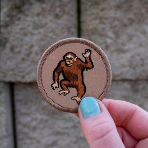 May include: A brown embroidered patch with a cartoon image of an orangutan with its arm raised in the air. The patch is circular and has a brown stitched border.