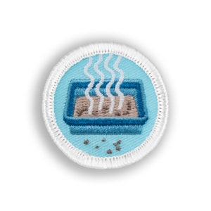 May include: A blue and white embroidered patch with a cartoon image of a litter box with brown cat litter and steam coming out of it. There are small brown dots scattered around the litter box.
