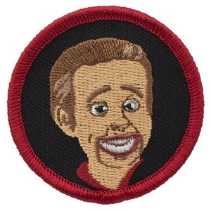 May include: Embroidered patch featuring a smiling cartoon man with brown hair, wearing a red shirt, against a black background with a red border.