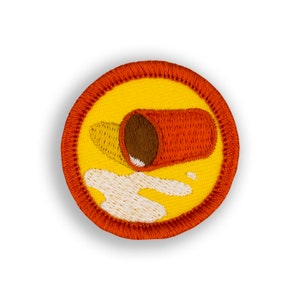 May include: A yellow and orange embroidered patch with a red and brown cup spilling white liquid. The cup is tipped over and the liquid is spilling out.