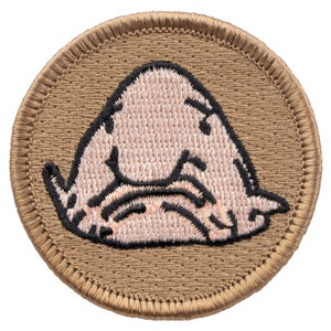 May include: A brown embroidered patch with a pink blobfish design. The blobfish is a deep-sea fish known for its unusual appearance.