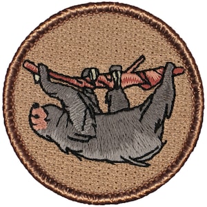 May include: A brown embroidered patch with a gray sloth hanging from a branch. The sloth is facing to the left and has a pink nose.