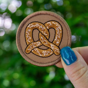 May include: A brown embroidered patch with a pretzel design. The pretzel is a light brown color with a white outline.