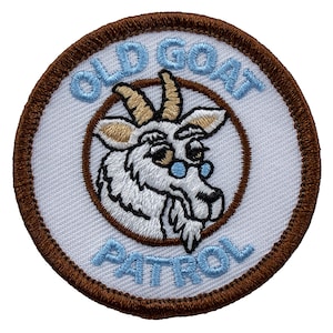 May include: A white embroidered patch with a brown border. The patch features a cartoon goat wearing glasses and the text "Old Goat Patrol".
