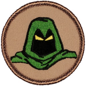 May include: A green hooded figure with yellow eyes on a brown background. The figure is embroidered on a round patch.