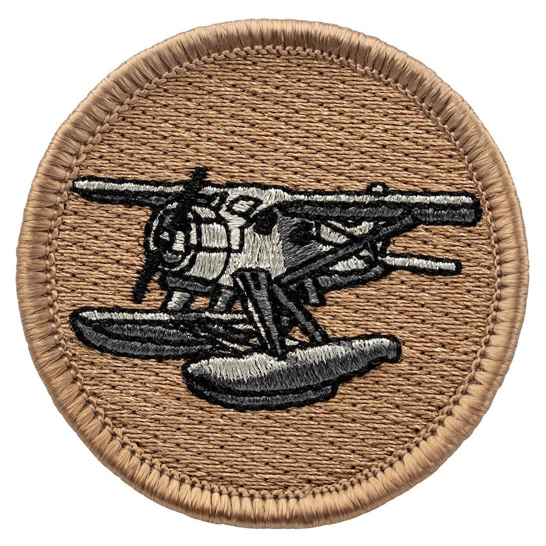 May include: A brown embroidered patch with a white and gray seaplane on it. The seaplane has two floats and is flying over water.