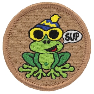 May include: A green frog wearing yellow sunglasses and a blue and yellow hat with a speech bubble that says "SUP". The frog is sitting on a brown background.