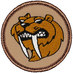 May include: A brown embroidered patch with a cartoon image of a saber-toothed animal with large white fangs. The patch is circular and has a brown stitched border.