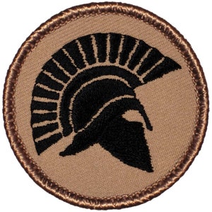 May include: Round patch featuring a black Spartan helmet silhouette on a tan background. The patch has a brown stitched border. The helmet design is detailed with vertical lines and a prominent cheek guard, creating a classic, historical aesthetic.