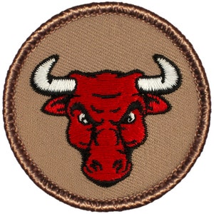 May include: A red embroidered bull's head with white horns on a brown background. The bull has a fierce expression with black eyes and a black nose.
