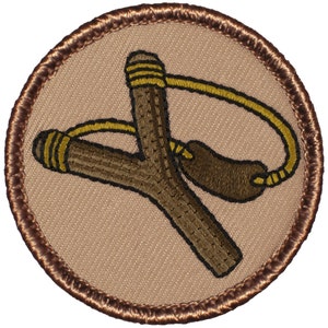 May include: A brown leather slingshot with a yellow rubber band. The slingshot is embroidered on a tan fabric patch with a brown border.