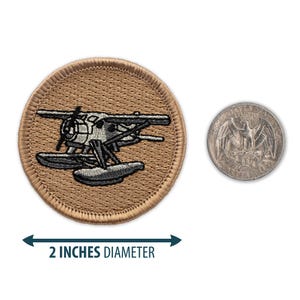 May include: A 2-inch diameter embroidered patch featuring a gray and white seaplane on a tan background. The patch is shown next to a US quarter for size comparison.