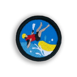 May include: A blue and yellow embroidered patch with a person in a red shirt and black shorts falling into the water. The person is holding a blue paddle and is being pulled by a yellow tube.