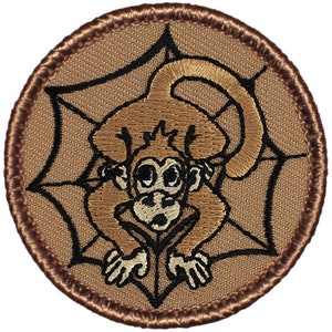 May include: A brown embroidered patch with a cartoon monkey in a spider web. The monkey is brown with black eyes and a black mouth. The spider web is black and the background is brown.