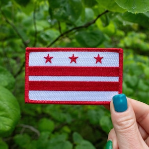 May include: Embroidered patch featuring the flag of Washington, D.C. The rectangular patch has a white background with three red stars at the top and two red horizontal stripes. The patch is bordered with a red stitch.