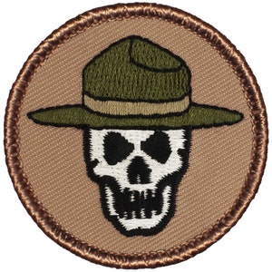 May include: A round, brown embroidered patch with a white skull wearing a green ranger hat. The patch has a brown border.
