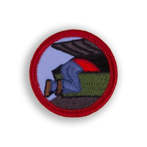 May include: Embroidered patch with a red border featuring a person in blue jeans and a red shirt, appearing to be climbing into a green box. The background is light blue and the top of the box is black.