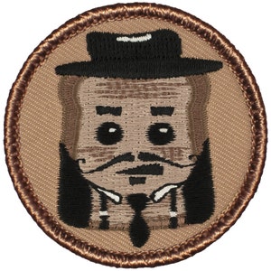 May include: Embroidered patch featuring a cartoon character with a brown face, black mustache, and black hat. The character is wearing a white shirt and a black tie. The patch is circular and has a brown border.