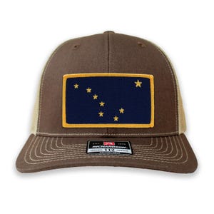 May include: A brown trucker hat with a tan mesh back and a patch featuring the Alaska state flag. The flag is a navy blue rectangle with gold stars and a gold border. The hat has a curved brim and an adjustable strap.